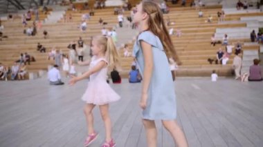 Mavi ve gül elbiseli iki sevimli uzun saçlı kız kardeş açık hava sahnesinde dans ediyor.