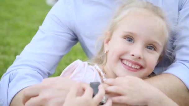 Pai abraça sua pequena filha adorável loira em rosa sentado no prado. close-up — Vídeo de Stock