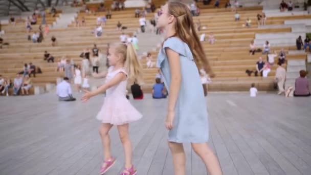 Twee schattige langharige zussen in blauw en roos jurken dansen op openlucht scène — Stockvideo