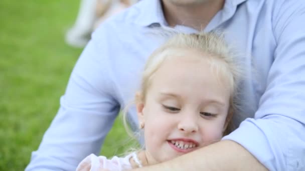 Papa knuffelt blonde dochter speels bijt zijn hand, zittend op weide. close-up — Stockvideo
