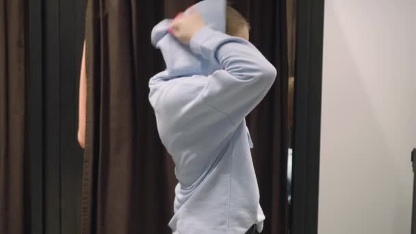 Mooie jonge vrouw probeert blauwe sweater, zet een kap over haar hoofd — Stockvideo