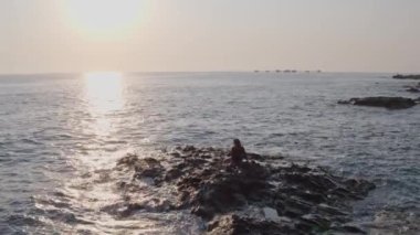 İHA uçuşu, kayalık bir tepede oturan bir kız, deniz manzaralı 4K