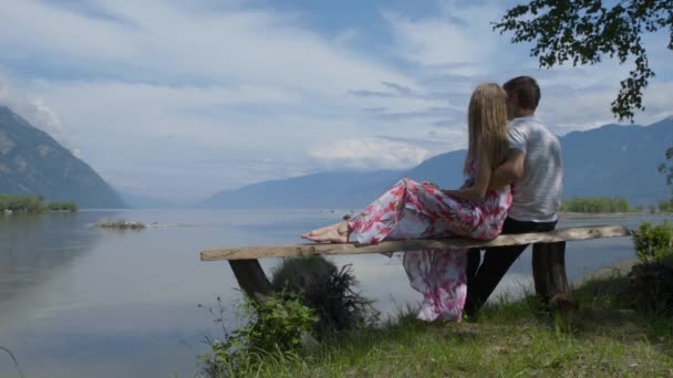Пара сидит на скамейке на берегу реки, разговаривает, смотрит на горы — стоковое видео