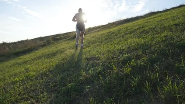 Молодий чоловік біжить над зеленим пагорбом на фоні блакитного неба. Чоловічий спортсмен бігає на природі під час заходу сонця. Спортивний бігун біжить вгору на світанку зі спалахом. Тренування між країнами. Повільний рух — стокове відео