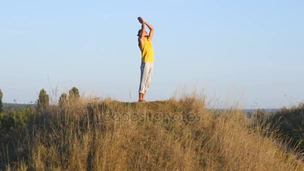 Hombre caucásico practicando movimientos de yoga y posiciones en la naturaleza. Joven deportista de pie en yoga posan al aire libre. Atleta balanceándose. Hermoso paisaje como fondo. Vida activa saludable — Vídeos de Stock