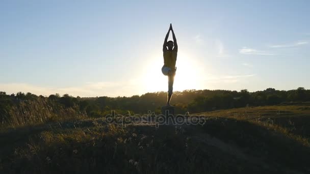 Yogui practicando movimientos de yoga y posiciones en la naturaleza. Silueta de hombre deportivo de pie en la pose de yoga al aire libre. Atleta balanceándose en una pierna. Hermoso cielo y sol como fondo. Vida activa saludable — Vídeo de stock