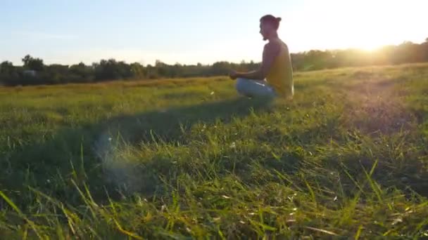 Молодой человек, сидящий в позе йоги у зеленой травы на лугу и медитирует. Мускулистый парень расслабляется в позе лотоса на природе. Отдых на открытом воздухе в солнечный день. Красивый пейзаж на заднем плане. Закрыть — стоковое видео