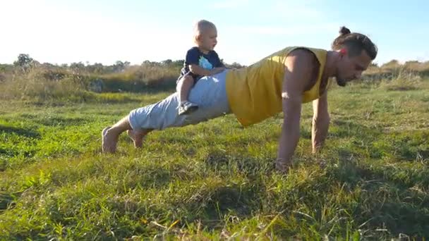 Молодой мускулистый отец отжимается с маленьким сыном на спине. Атлетик отжимается на зеленой траве с счастливым улыбающимся ребенком, сидящим на заднице. Сильный парень проводит время с вашим ребенком — стоковое видео
