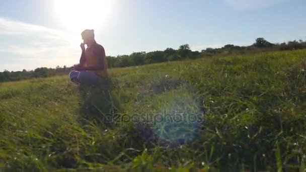 Młody człowiek siedzi w jodze stanowią o zielonej trawy na łące i medytuje. Muskularny facet na relaks w pozycji lotosu w nature. Rekreacja na świeżym powietrzu w słoneczny dzień. Piękny krajobraz w tle. Z bliska — Wideo stockowe