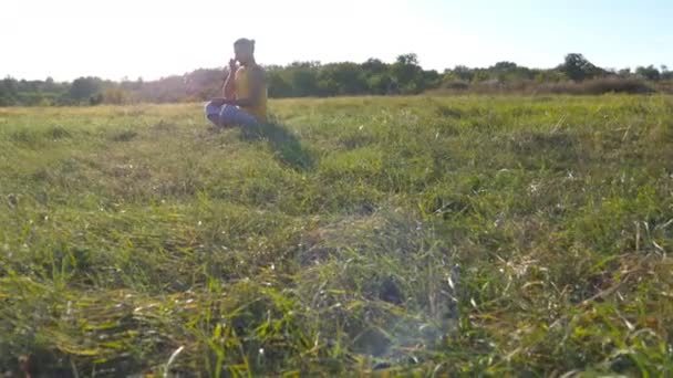 Jonge man zitten in yoga vormen op groen gras in de wei en mediteert. Gespierde man ontspannen in lotus houding in de natuur. Recreatie buiten op een zonnige dag. Mooi landschap op de achtergrond. Close-up — Stockvideo