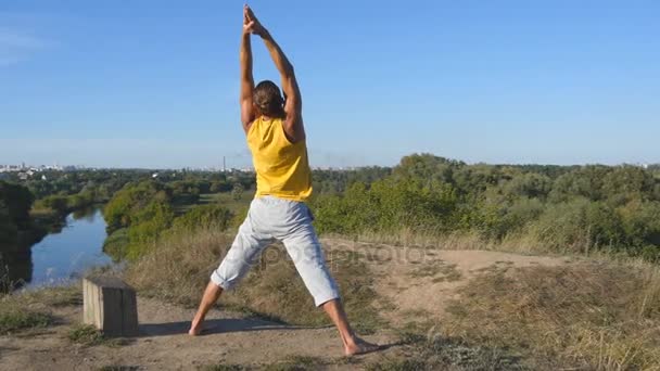 Joven parado en el yoga posan en la naturaleza. Chico practicando movimientos de yoga y posiciones al aire libre. Atleta haciendo ejercicio de fuerza en la colina. Paisaje de fondo. Vida activa saludable. De cerca. — Vídeo de stock