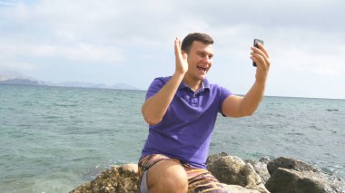 Video aramasý deniz plajda akıllı telefon olan genç gülümseyen adam. Mutlu adam video çevrimiçi sohbetler güzel manzara arka plan yapma. Çocuk cep ve gösteren okyanus üzerinde konuşuyor. Selfie yapan erkek