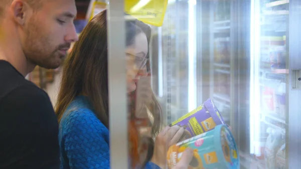 Молодая счастливая пара стоит у холодильника в продуктовом магазине и собирает пакеты с замороженной едой. Пара забирает товар из холодильника и кладет его в корзину. Выбор мороженого в магазине . — стоковое фото
