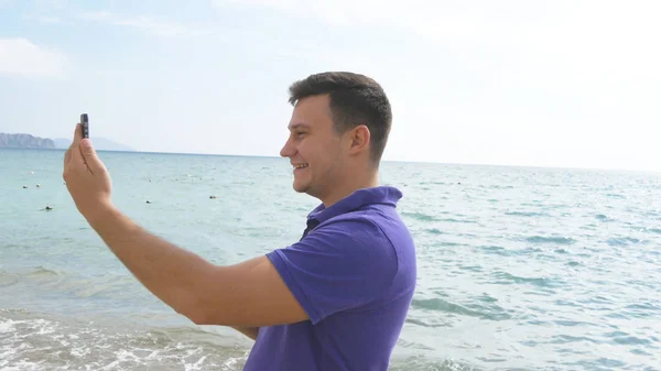 Junger lächelnder Mann bei einem Videoanruf auf dem Smartphone am Strand. Glücklicher Kerl, der Online-Videochats auf wunderschönem Landschaftshintergrund macht. Junge spricht auf Handy und zeigt Ozean. Mann macht Selfie — Stockfoto