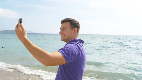 Junger lächelnder Mann bei einem Videoanruf auf dem Smartphone am Strand. Glücklicher Kerl, der Online-Videochats auf wunderschönem Landschaftshintergrund macht. Junge spricht auf Handy und zeigt Ozean. Mann macht Selfie — Stockfoto