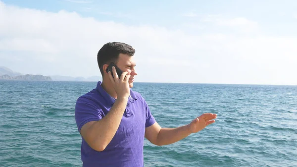 Ein junger, gutaussehender Mann telefoniert am Strand. Seriöser Typ, der auf dem Handy vor dem Hintergrund des Ozeans spricht. Anruf aus den Sommerferien. schöne Landschaft. Nahaufnahme — Stockfoto