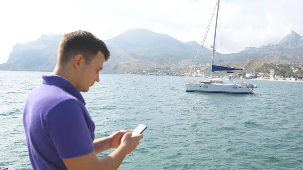 Junger Mann, der am Meer steht und sein Smartphone benutzt. Jacht im Meer im Hintergrund. Porträt eines gutaussehenden Typen mit Handy am Strand. SMS schreiben oder in sozialen Netzwerken chatten. Nahaufnahme — Stockfoto