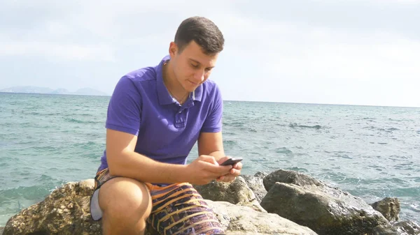 Jongeman is zittend op een steen en het gebruik van smartphone in de buurt van de zee. Portret van knappe jongen met mobiele telefoon op het strand van de Oceaan. Jongen telefoon app kijken. Hij SMS sms of chatten in sociaal netwerk — Stockfoto