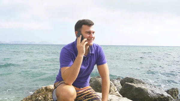 Ein junger lächelnder Mann telefoniert am Strand im Meer. gut aussehender glücklicher Kerl, der auf Steinen in der Nähe des Meeres sitzt und auf einem Handy mit wunderschönem Landschaftshintergrund spricht. Nahaufnahme — Stockfoto