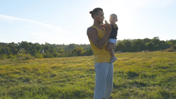 Młody ojciec trzyma w ręce jego syna na łące w nature w słoneczny letni dzień. Tata i mały chłopiec, przytulanie odkryty i dając pięciu do siebie. Piękny krajobraz w tle. Z bliska — Zdjęcie stockowe
