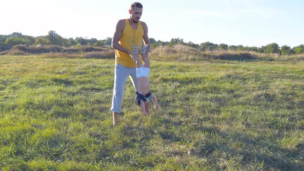 Joven padre jugando con su hijo en la naturaleza. Papá sostiene las piernas de sus hijos y las lanza al aire libre. Feliz familia pasando tiempo juntos afuera en el prado. Hermoso paisaje de fondo. De cerca. — Foto de Stock