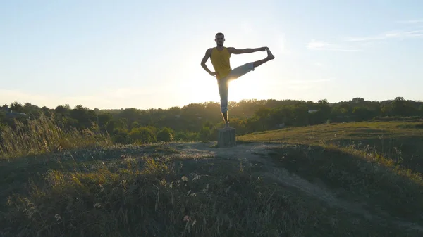 Egy sportos férfi sziluettje áll a jóga pózon a szabadban. Jóga gyakorló jóga mozdulatok és pozíciók a természetben. A sportoló az egyik lábán egyensúlyozik. Gyönyörű ég és nap, mint háttér. Egészséges aktív életmód — Stock Fotó