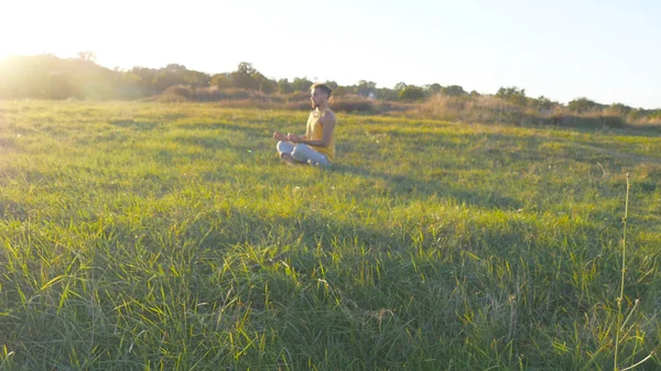 Young man sitting in yoga pose at green grass in the meadow and meditates. Muscular guy relaxing in lotus pose at nature. Recreation outdoor at sunny day. Beautiful landscape at background. Close up