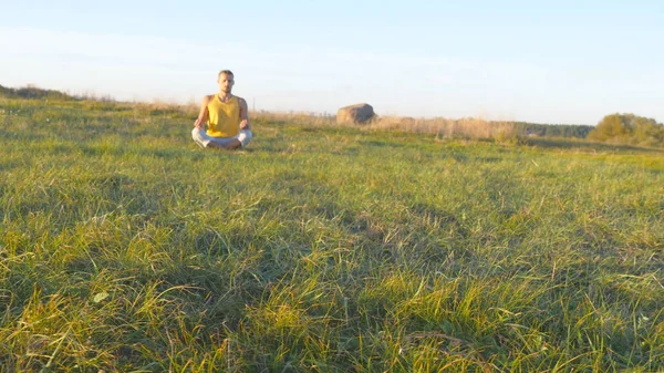 Giovane seduto in posa yoga a erba verde nel prato e medita. Il tipo muscolare che si rilassa in loto posa a natura. Attività ricreative all'aperto nella giornata di sole. Bellissimo paesaggio sullo sfondo. Da vicino. — Foto Stock
