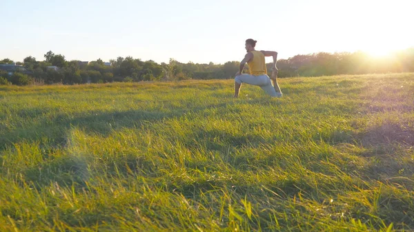 Młody człowiek siedzący na zielonej trawie w łące i robi ćwiczenia jogi. Muskularny facet, rozciągając jego ciało w nature. Szkolenia na zewnątrz w słoneczny dzień. Piękny krajobraz w tle. Z bliska — Zdjęcie stockowe