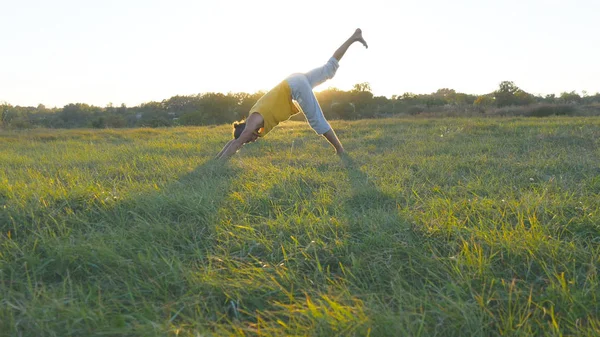 Młody człowiek praktykowania jogi przenosi i pozycje o zielonej trawy na łące. Sportowy facet stojący w jogi w przyrodzie. Piękny krajobraz w tle. Zdrowy, aktywny tryb życia jogina odkryty — Zdjęcie stockowe