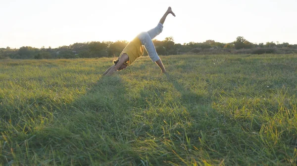 Joven practicando yoga se mueve y posiciona en la hierba verde en el prado. Tipo deportivo de pie en la pose de yoga en la naturaleza. Hermoso paisaje como fondo. Vida activa saludable del yogui al aire libre —  Fotos de Stock