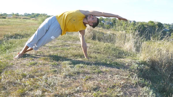 En fyr, der øver yogabevægelser og stillinger udendørs. Ung mand, der står ved yoga, poserer ved naturen. Atleten laver styrketræning på bakken. Landskab på baggrund. En sund og aktiv livsstil. Tæt på . - Stock-foto