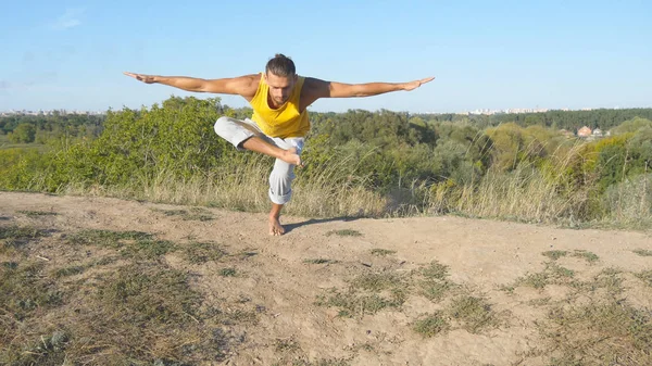Sportos fiatalember állt fa jóga póz szabadtéri. Kaukázusi srác jóga mozog, és a természetben pozíciók. Yogi kiegyensúlyozó egyik lábát. Gyönyörű táj, mint háttér. Egészséges és aktív életmód — Stock Fotó