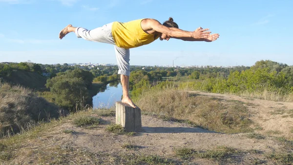 Ung sporty mand stående ved yoga positur udendørs. Kaukasiske fyr praktiserer yoga bevægelser og positioner i naturen. Atleten balancerer på et ben. Smukt landskab som baggrund. Sund aktiv livsstil - Stock-foto