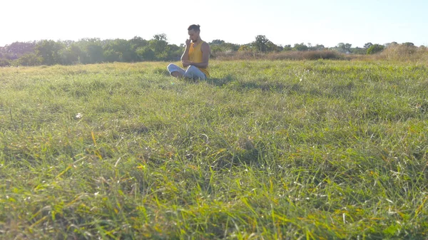 ヨガで座っている若い男は、緑の草の牧草地でポーズし、瞑想します。筋肉男は自然でロータスのポーズでリラックス。晴れた日の屋外のレクリエーション。バック グラウンドで美しい風景。クローズ アップ — ストック写真