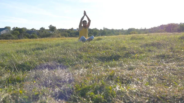 Young man sitting in yoga pose at green grass in the meadow and meditates. Muscular guy relaxing in lotus pose at nature. Recreation outdoor at sunny day. Beautiful landscape at background. Close up