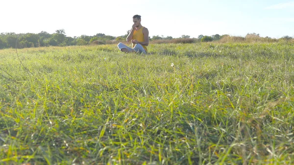 Młody człowiek siedzi w jodze stanowią o zielonej trawy na łące i medytuje. Muskularny facet na relaks w pozycji lotosu w nature. Rekreacja na świeżym powietrzu w słoneczny dzień. Piękny krajobraz w tle. Z bliska — Zdjęcie stockowe