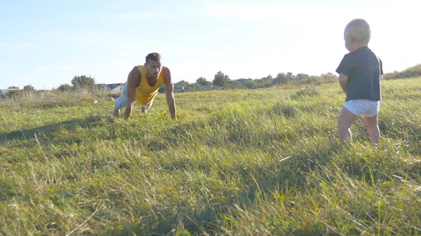 Pequeño bebé de pie sobre hierba verde en el prado y mirando como su padre haciendo flexiones. Hombre atlético haciendo flexiones en la naturaleza, hijo mirándolo. Fuerte padre pasar tiempo con su hijo al aire libre . — Foto de Stock