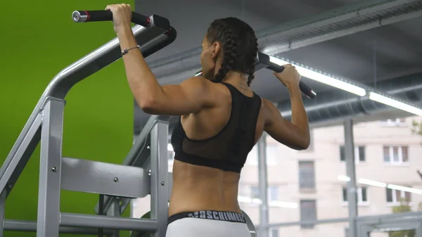 Chica de fitness haciendo flexiones en el gimnasio. Mujer joven haciendo ejercicios en la barra horizontal. Entrenamiento físico. Vista trasera trasera — Foto de Stock