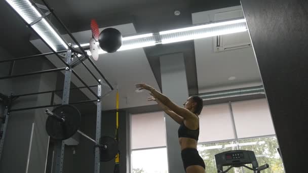 Mujer joven y fuerte con un cuerpo de fitness perfecto en ropa deportiva haciendo ejercicio con balón de medicina en el gimnasio. Chica haciendo entrenamiento crossfit. Atleta hembra sentadillas durante el entrenamiento en interiores. En cámara lenta. De cerca. — Vídeo de stock