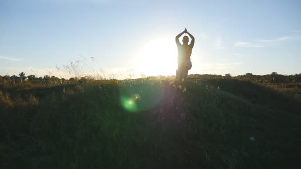 Młody człowiek praktykowania jogi przenosi i pozycje na wzgórzu o zachodzie słońca. Sportowy facet stojący w jogi w przyrodzie. Piękny krajobraz w tle. Zdrowy, aktywny tryb życia jogina odkryty. Zwolnionym tempie — Wideo stockowe