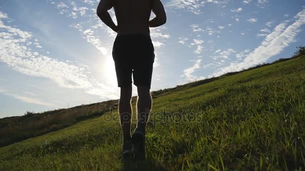 Mladý muž přejel zelený kopec na pozadí modré oblohy. Sportovec je běhat v přírodě při západu slunce. Sportovní běžce běhat do kopce venkovní za úsvitu s hrdlem. Běžecké školení. Zpomalený pohyb