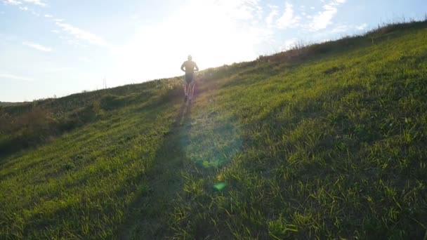 Ung man kör över grön kulle över blå himmel bakgrund. Manliga idrottare jogging i naturen vid solnedgången. Sport löpare jogging uppförsbacke utomhus vid soluppgången med flare. Cross-Country utbildning. Slow motion — Stockvideo