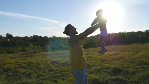 Молодой отец играет со своим маленьким сыном на открытом воздухе. Папа поднимает своего ребенка на природе. Счастливая семья проводит время вместе на лугу. Красивый пейзаж на заднем плане. Закрыть — стоковое видео