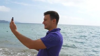 Video aramasý deniz plajda akıllı telefon olan genç gülümseyen adam. Mutlu adam video çevrimiçi sohbetler güzel manzara arka plan yapma. Çocuk cep ve gösteren okyanus üzerinde konuşuyor. Selfie yapan erkek