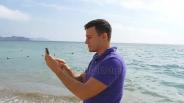 Video aramasý deniz plajda akıllı telefon olan genç gülümseyen adam. Mutlu adam video çevrimiçi sohbetler güzel manzara arka plan yapma. Çocuk cep ve gösteren okyanus üzerinde konuşuyor. Selfie yapan erkek