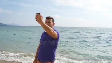 Video aramasý deniz plajda akıllı telefon olan genç gülümseyen adam. Mutlu adam video çevrimiçi sohbetler güzel manzara arka plan yapma. Çocuk cep ve gösteren okyanus üzerinde konuşuyor. Selfie yapan erkek