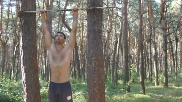 Atletische gespierde man met perfecte fitness lichaam uitvoeren van buikspieroefeningen op de horizontale balk aan het forest. Man heffen van de benen. Guy opleiding in het hout buiten. Hij opknoping en verhogingen bezinksel. — Stockvideo