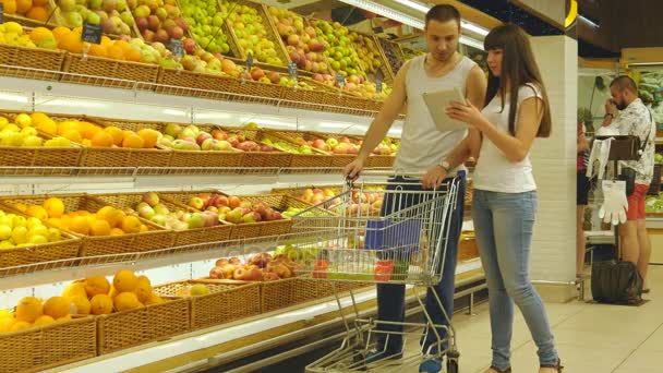Молодая кавказская пара, прогуливающаяся в супермаркете с тележкой и выбирающая свежие яблоки. Человек кладет фрукты в корзину магазина. Женщина использует планшетный ПК, чтобы проверить список покупок — стоковое видео