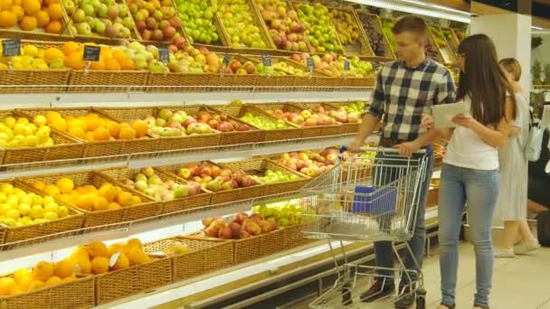 Jovem casal caucasiano caminhando em um supermercado com um carrinho de mercado e escolhendo maçãs frescas. O homem está a pôr frutos no cesto da loja. Mulher está usando tablet pc para verificar lista de compras — Vídeo de Stock